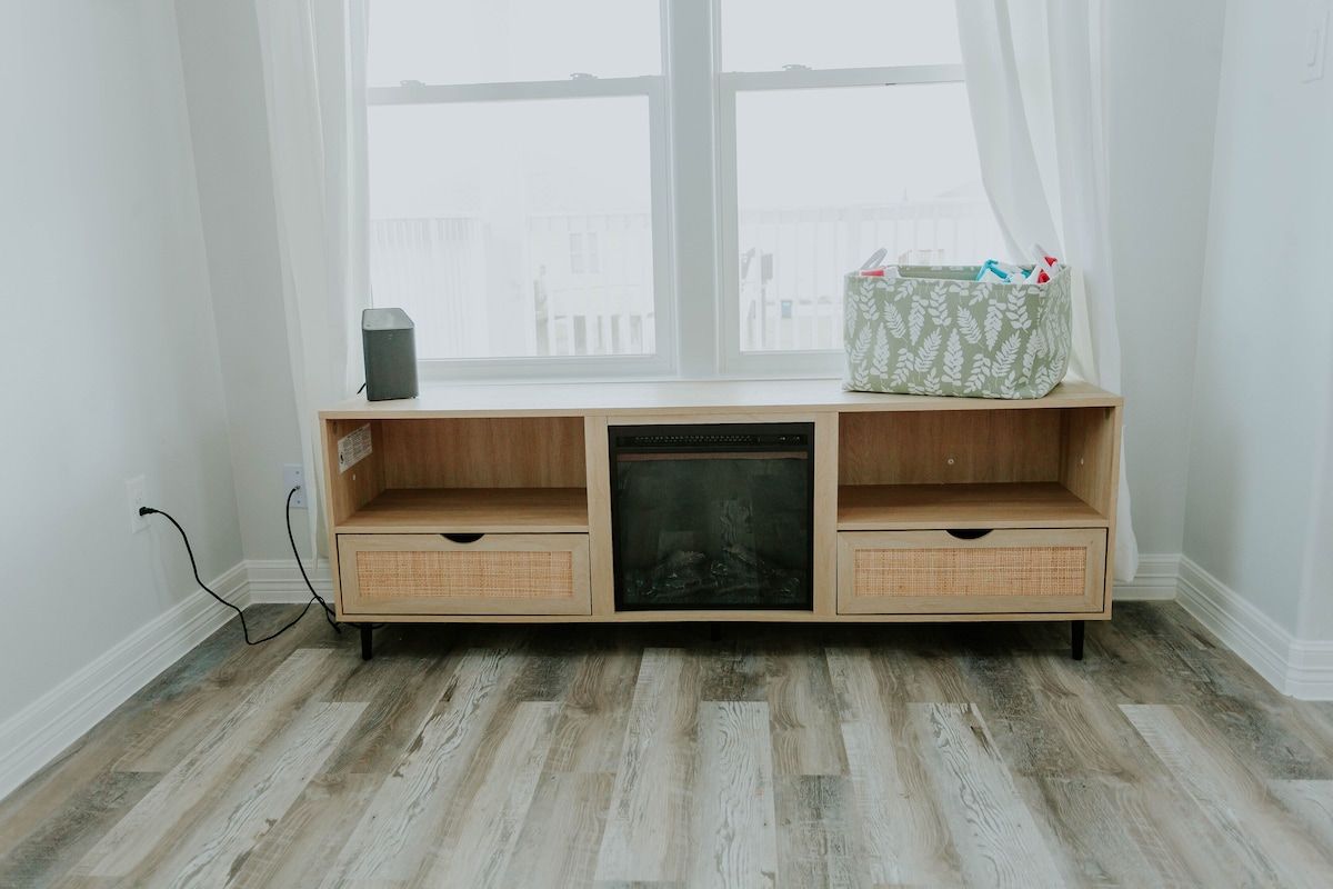 Light-wood media console with an integrated fireplace, two drawers, and open shelving, placed against a window.