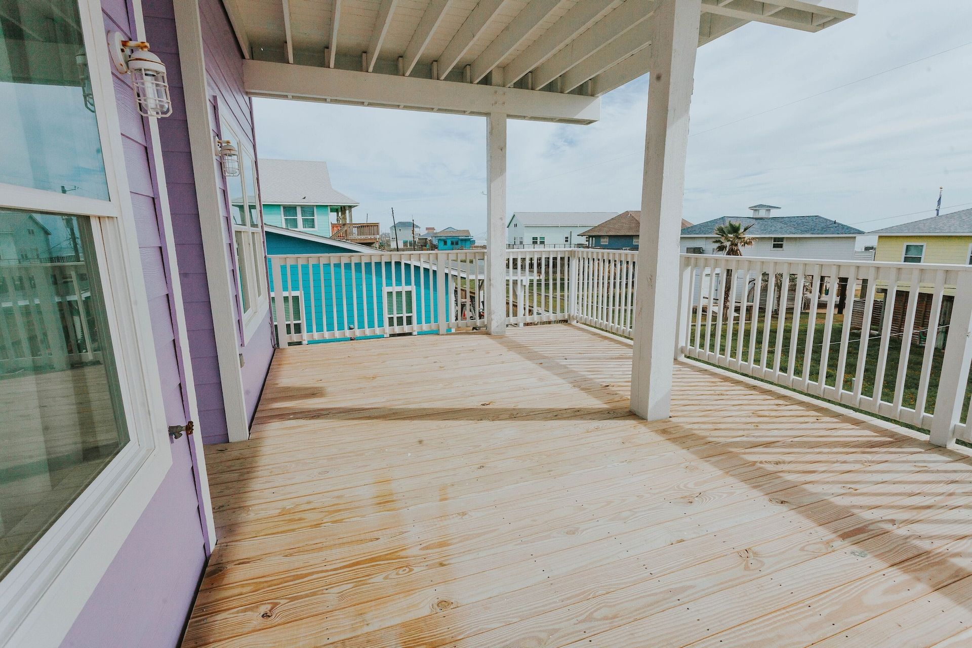 A light wooden deck with white railings under a covered patio, overlooking a residential beach area with colorful homes.