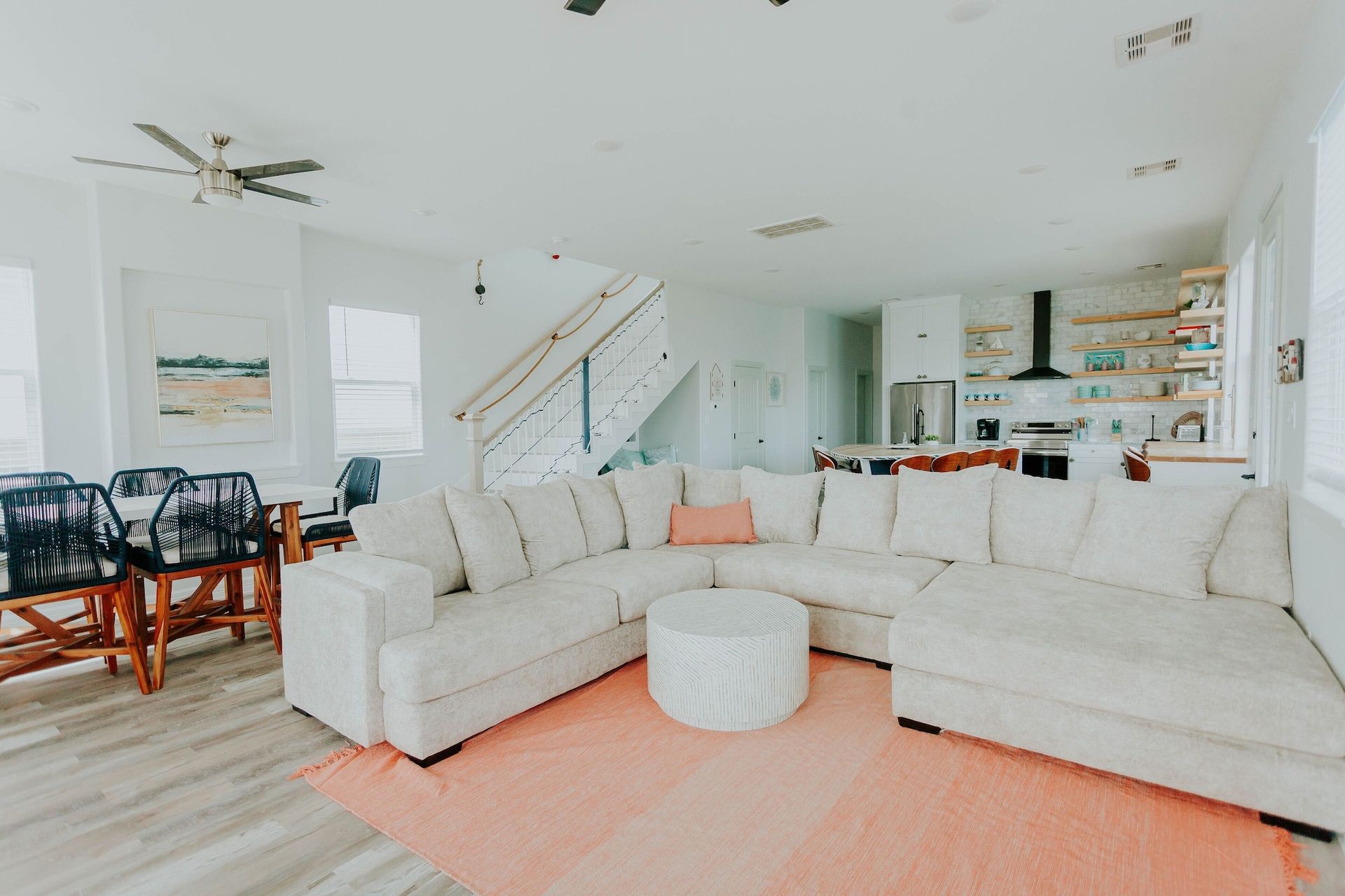 A bright, open-concept living and dining area with a large beige sectional, a peach rug, and a kitchen in the background.