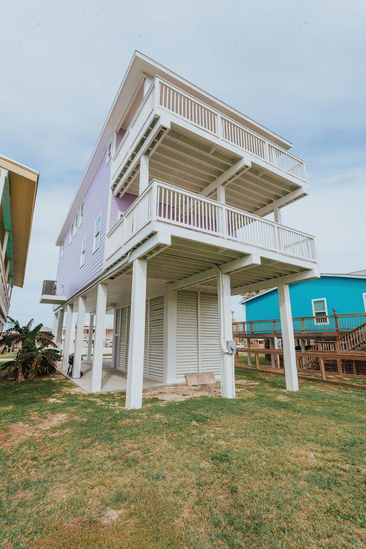 A tall, light-purple, three-story beach house elevated on white stilts above a grassy yard under a cloudy sky.