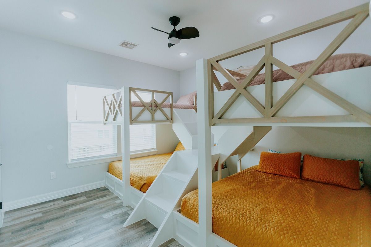 A bedroom with two white bunk beds, mustard yellow bedding, orange pillows, and light wood flooring.