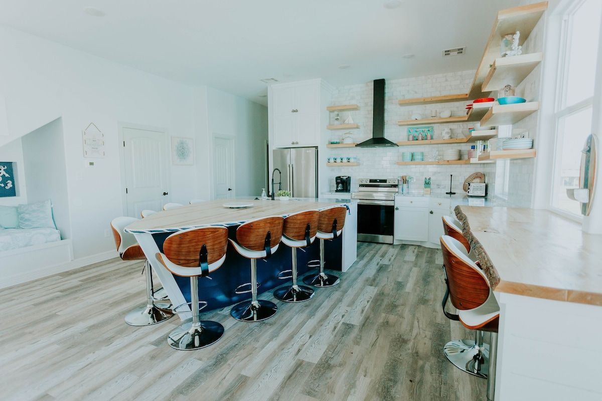 A modern, open-concept kitchen with a large central island, barstools, wood shelving, white cabinets, and wood-look flooring.