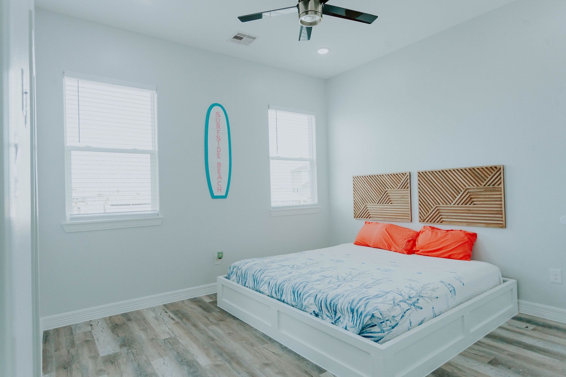 A bedroom with light blue walls, wood-look flooring, a white platform bed with orange pillows, and wall-mounted surf decor.