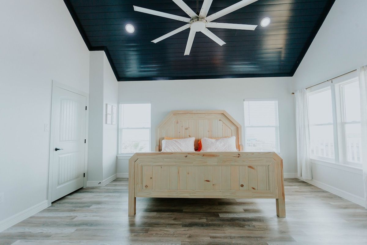 A modern bedroom with light wood flooring, a wooden bed frame, white walls, and a vaulted ceiling painted dark navy blue.