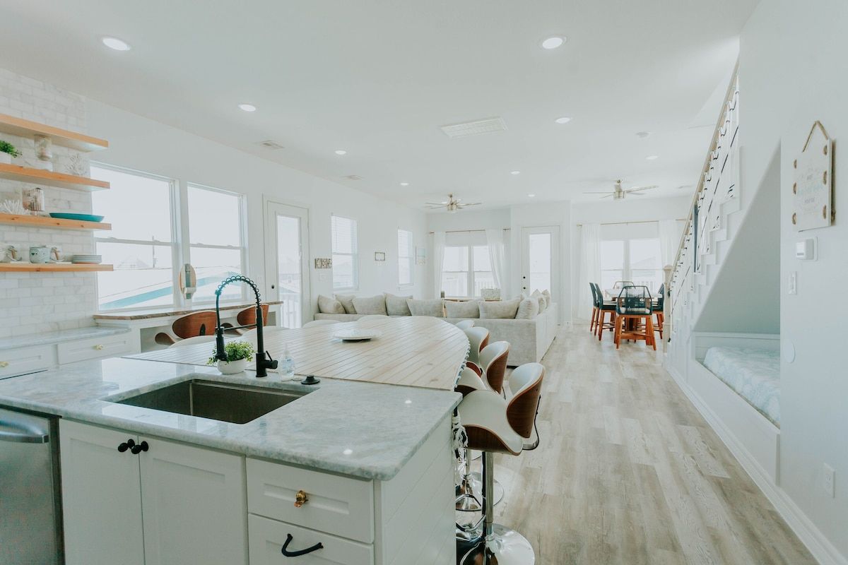 A bright, open-concept kitchen and living area with white cabinets, a marble island, wood flooring, and stairs.