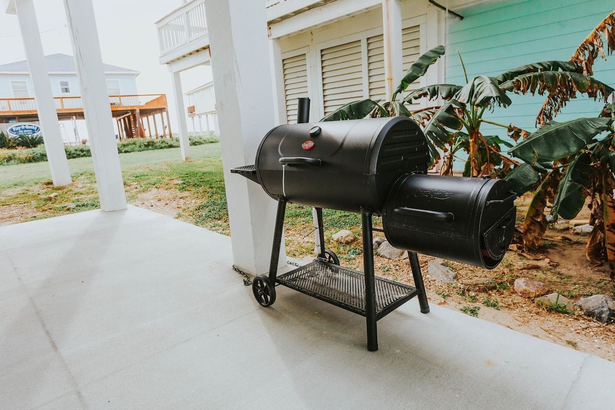 A black offset smoker grill sits on a concrete patio beside white house pillars and leafy green plants.