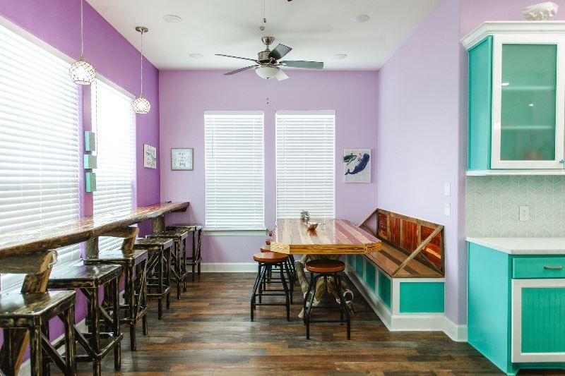 A kitchen with purple walls , a wooden table , stools , and a ceiling fan.