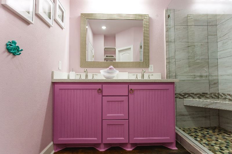 A bathroom with pink cabinets , a mirror , and a walk in shower.