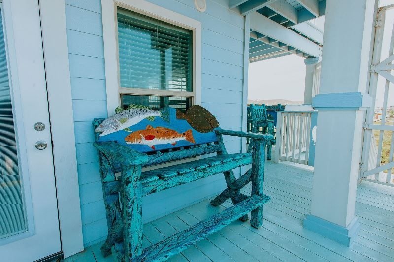 A blue bench with a fish on it is on a porch next to a window.