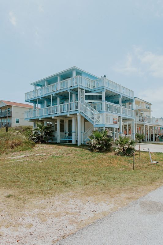 A large blue house with a lot of balconies and stairs is sitting on top of a grassy hill.