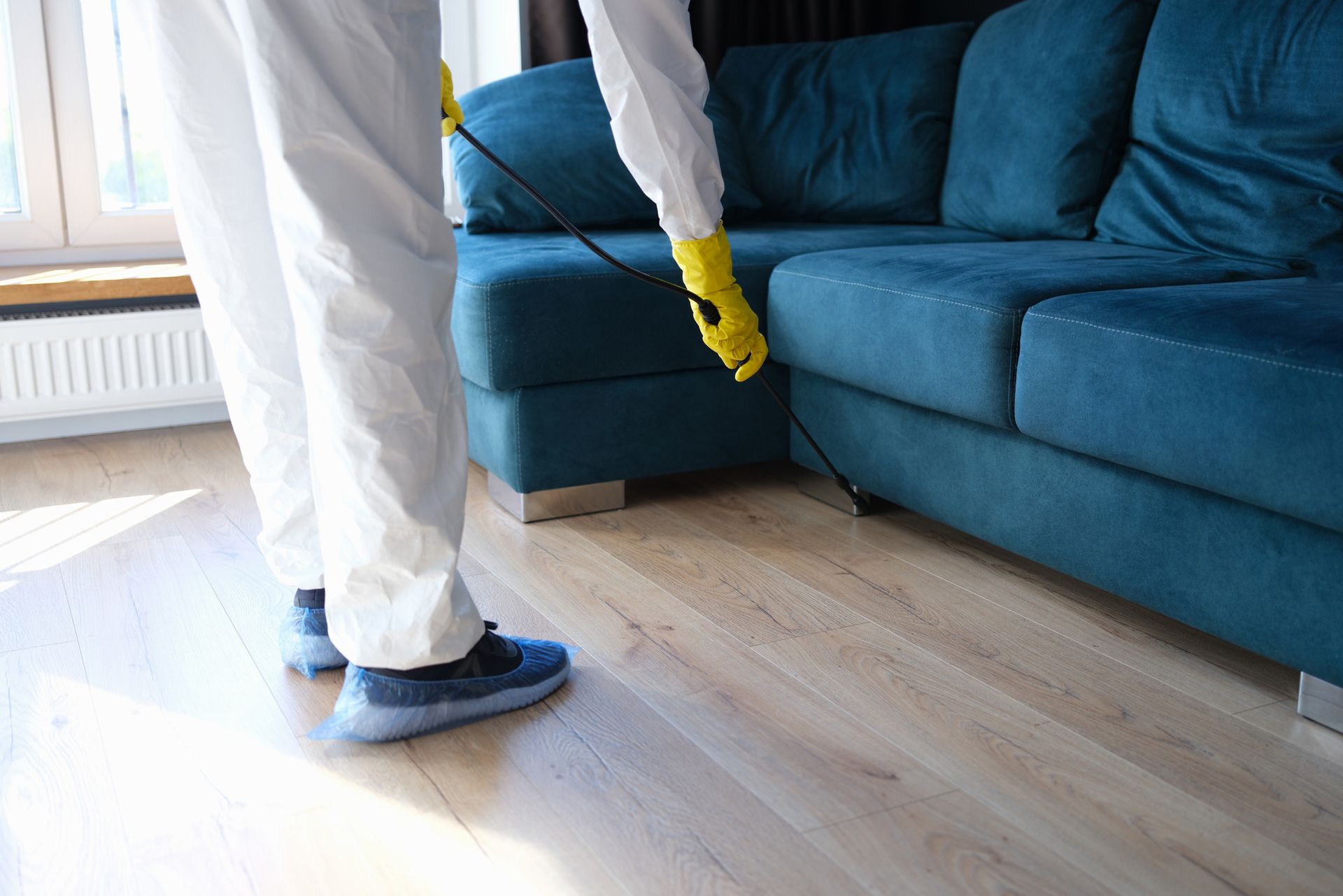 A pest control contractor, wearing safety gear, sprays under a green sofa, inside a living room.