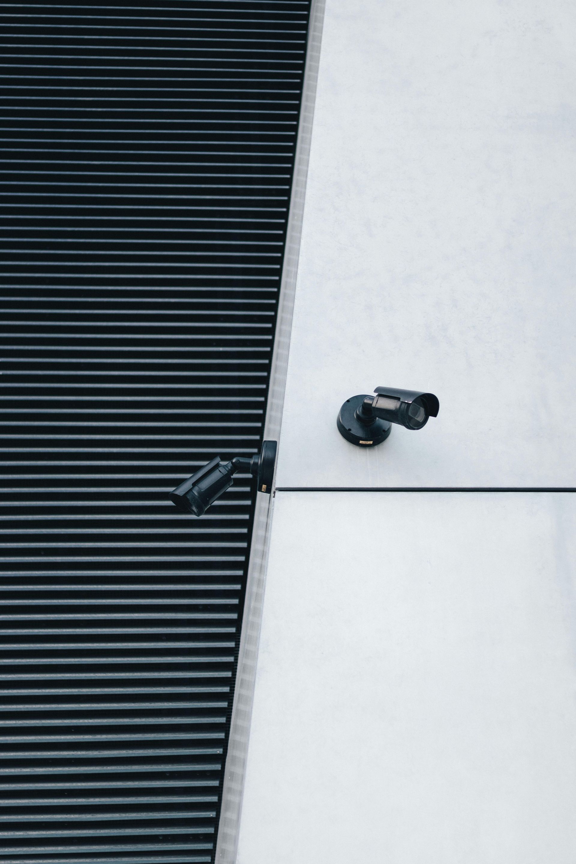 Two security cameras mounted on a white wall next to a dark, slatted wall.