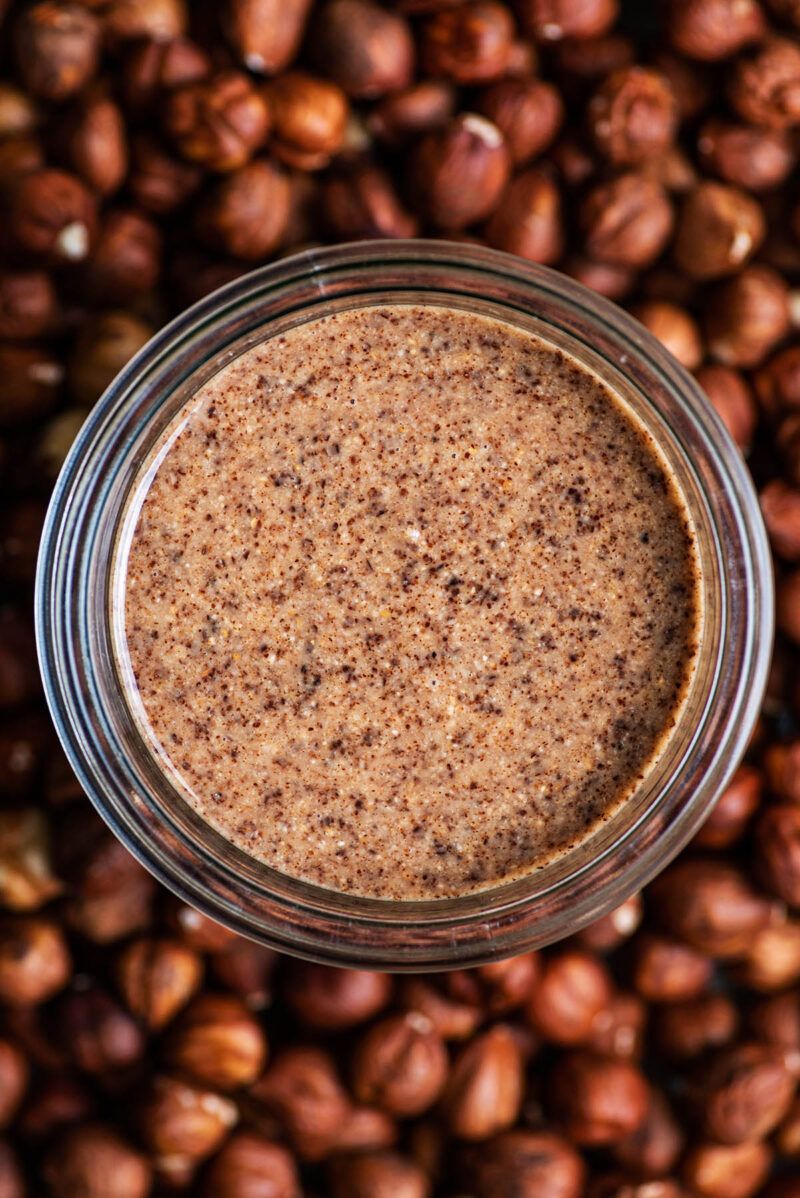 A jar of hazelnut butter is sitting on top of a pile of hazelnuts.
