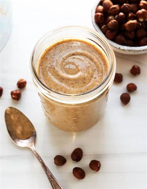 A jar of peanut butter next to a spoon and a bowl of peanuts.