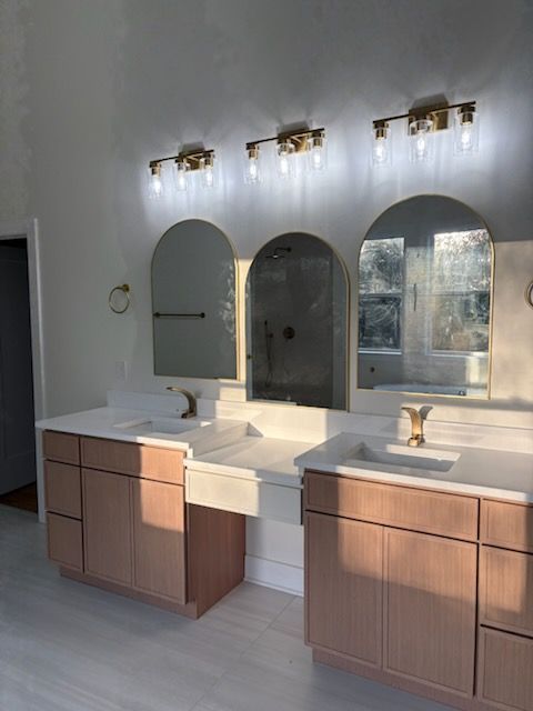 Bathroom with three arched mirrors, pink cabinets, gold fixtures, and light-colored flooring.