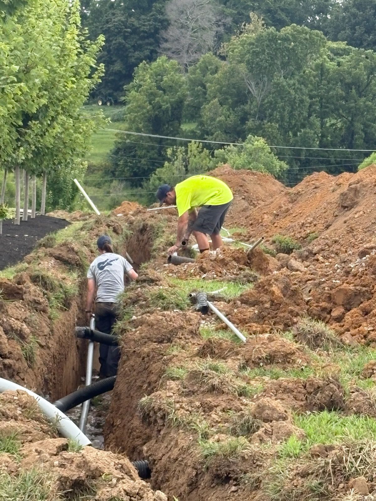 Two workers installing pipes in a trench outdoors. One in neon yellow, the other in gray.