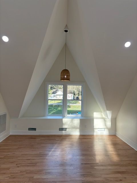 Empty room with wood floors, a triangular vaulted ceiling, and a window. A hanging light fixture is centered.