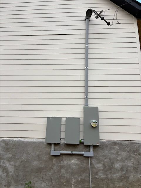 Electrical meter and breaker boxes mounted on a beige building exterior. Gray conduit runs up to overhead wires.