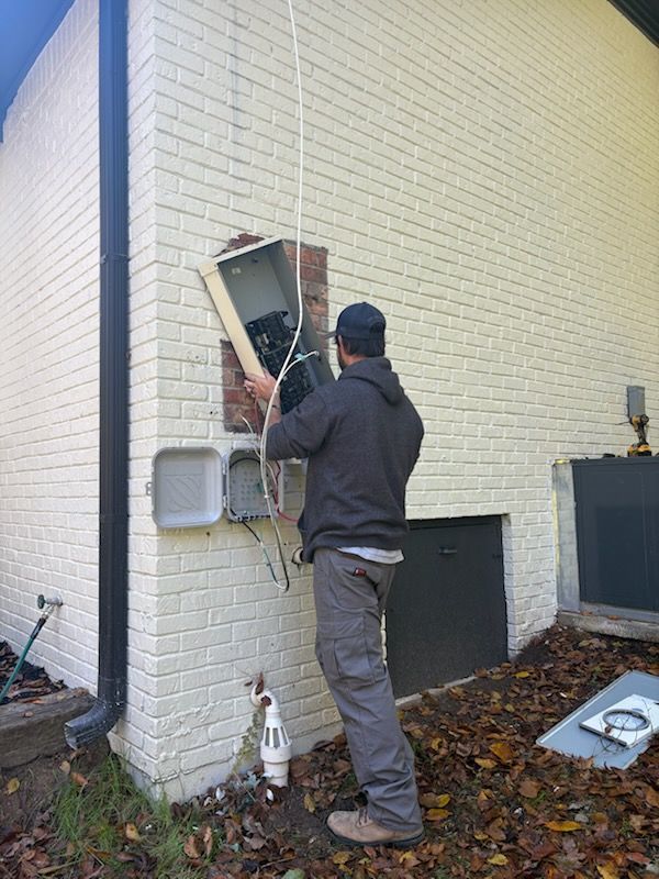 Man working on electrical box mounted on a brick wall. Wires hang down. Outdoors, fall foliage present.
