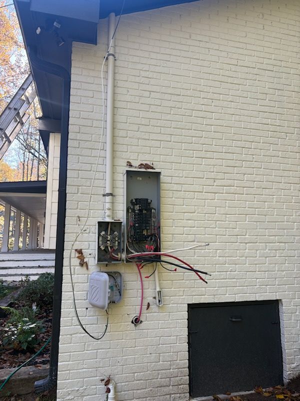 Exterior electrical panel on a brick wall, with exposed wires and a conduit running up.
