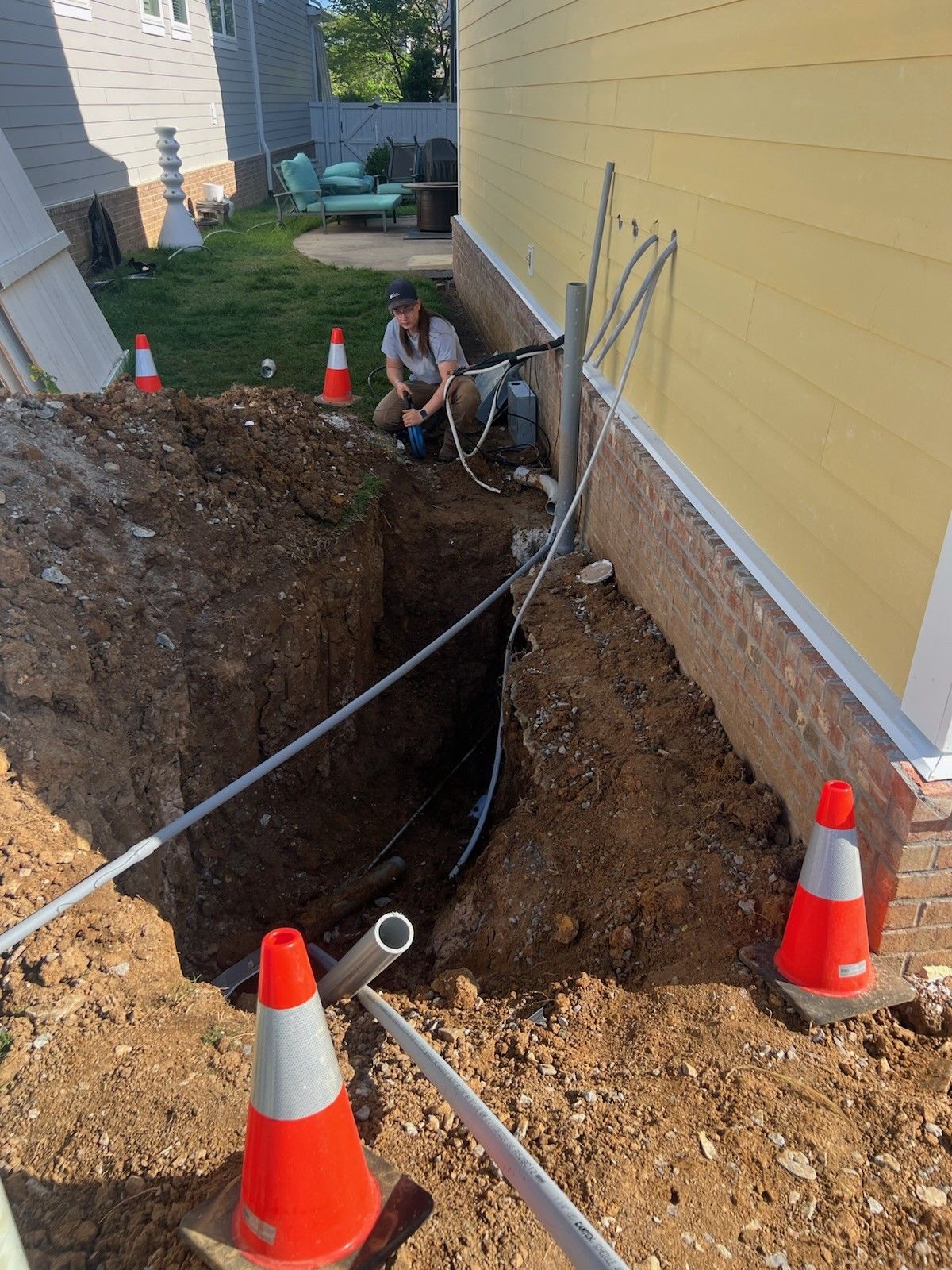 Person working on electrical lines in a trench next to a yellow house; orange cones mark the work area.