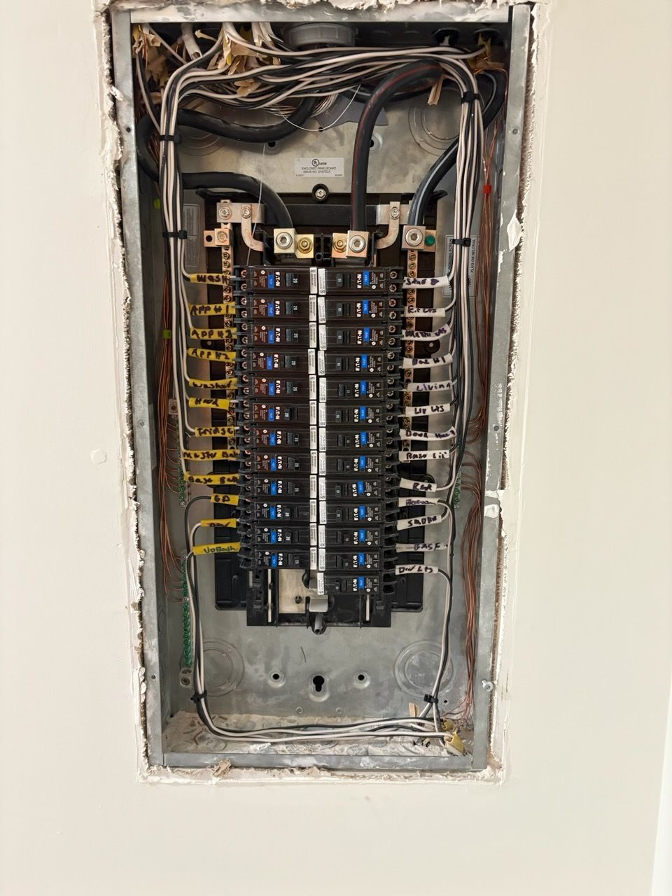 Electrical panel with numerous circuit breakers and wires inside a wall opening.