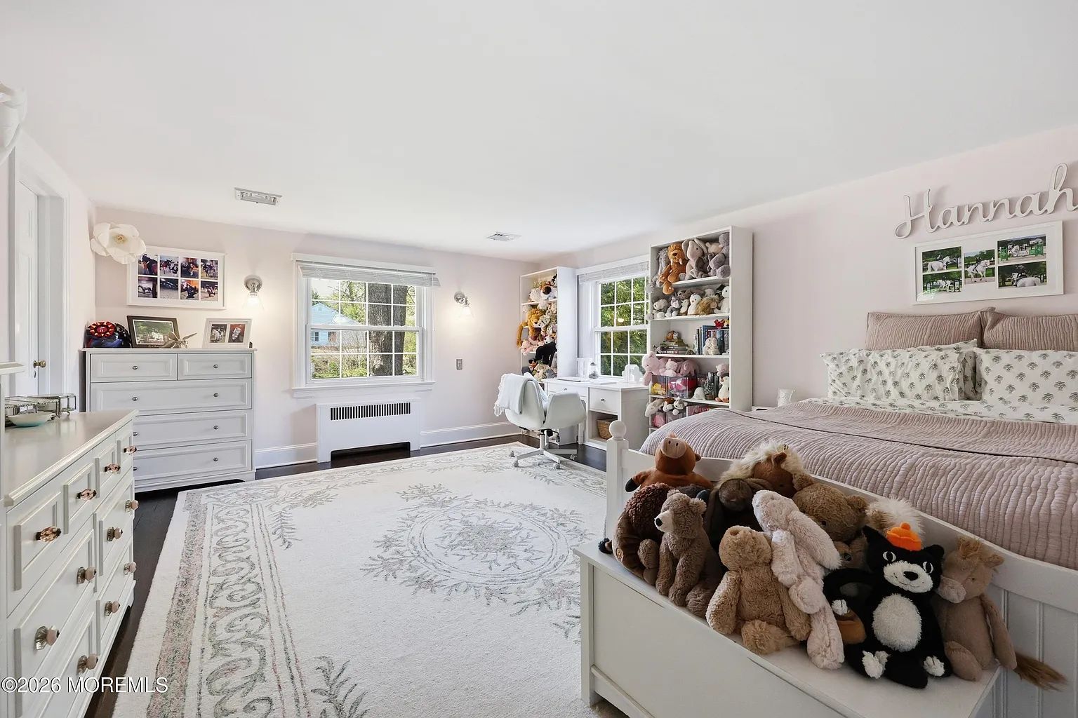 Bright white bedroom with pink accents, a bed, dresser, toys, and a window with plants and wall art.