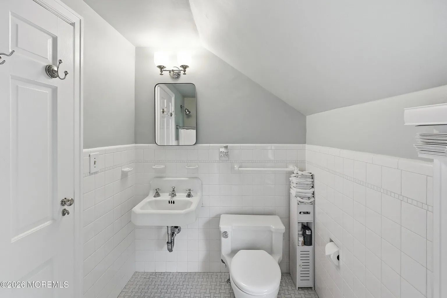 Bright white bathroom with sink, toilet, mirror, and tiled walls in a sloped-ceiling room