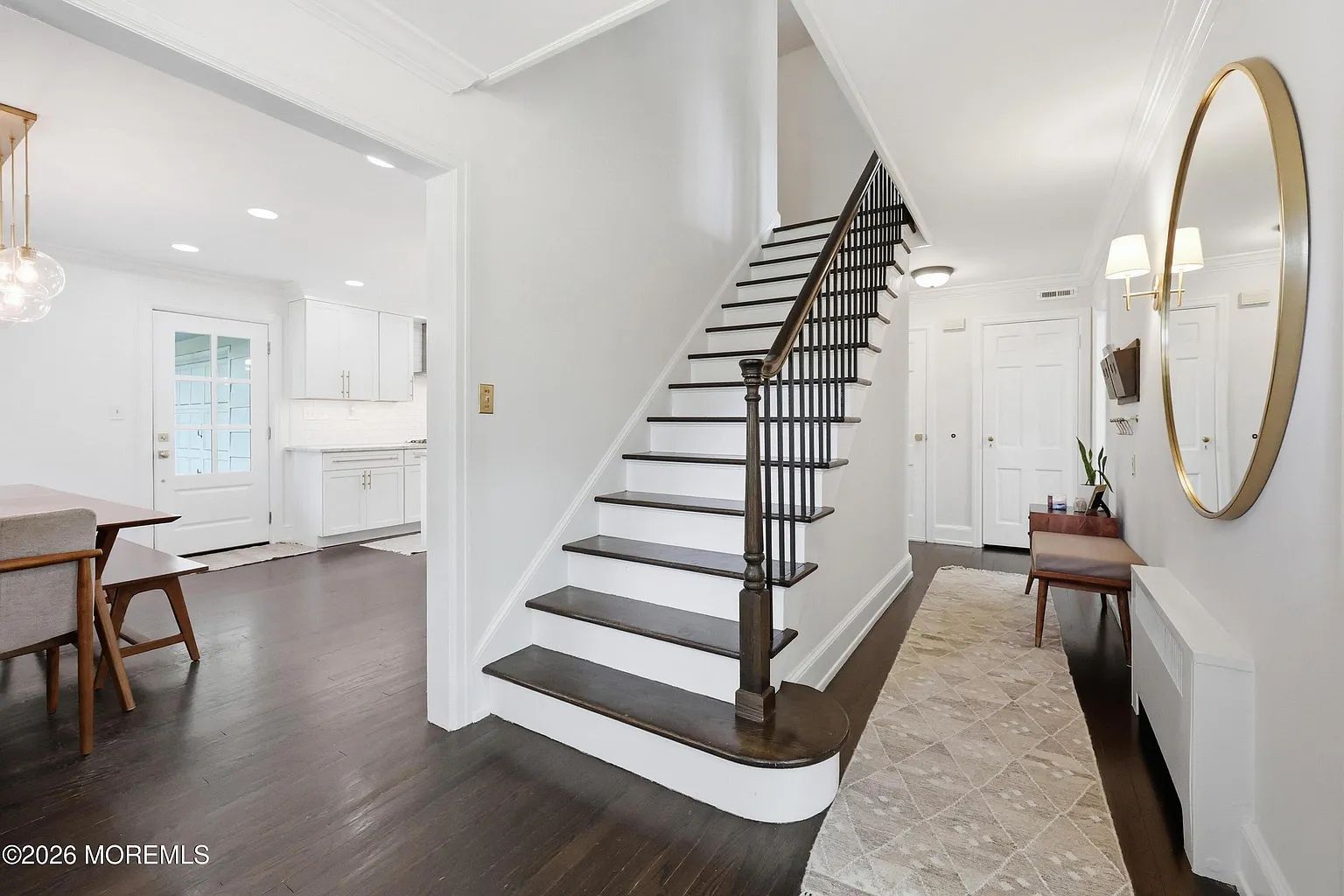 Bright foyer with dark staircase, hardwood floors, round mirror, and adjacent white kitchen and dining area.