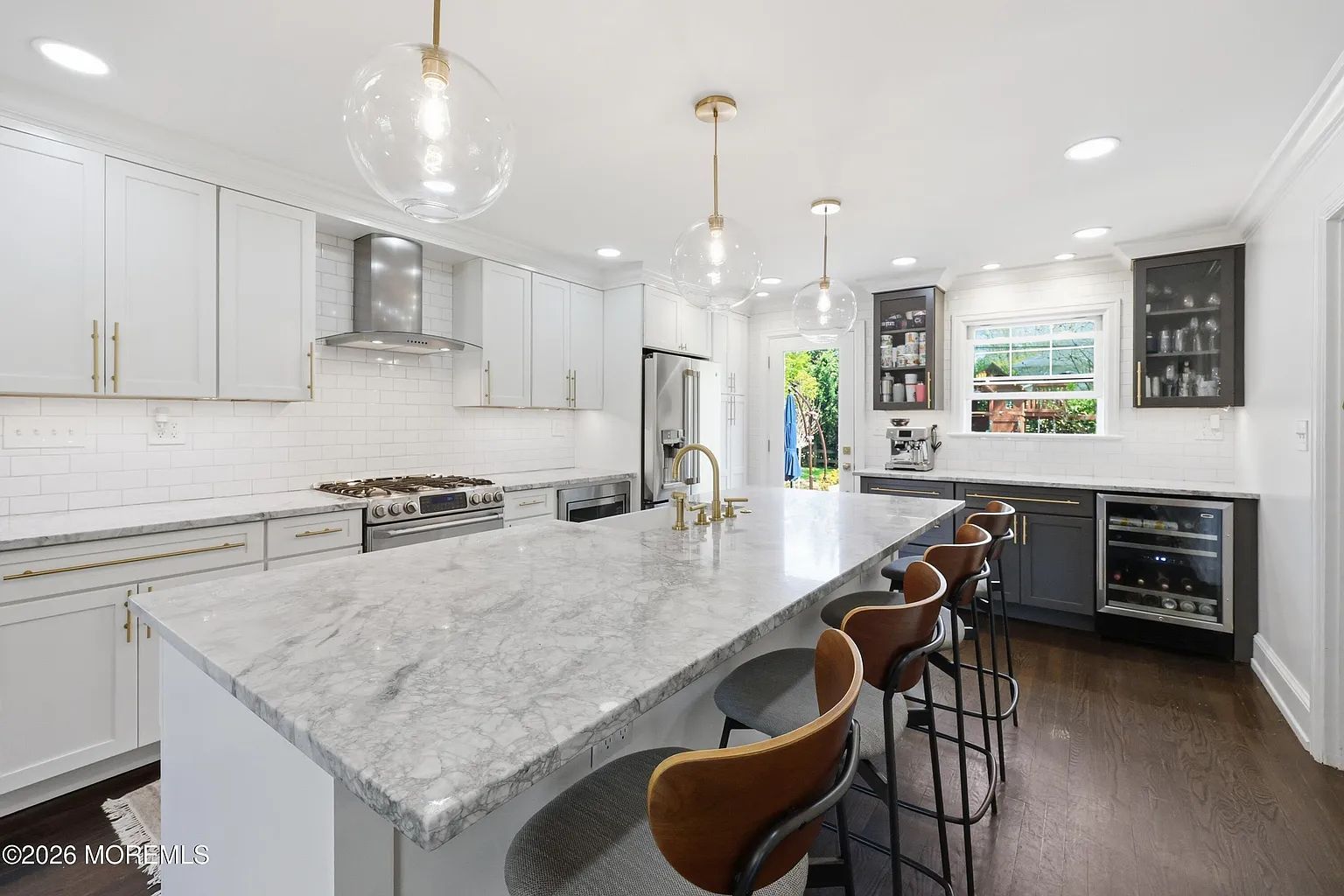 Bright modern white kitchen with marble island, bar stools, stainless appliances, and dark wood floors