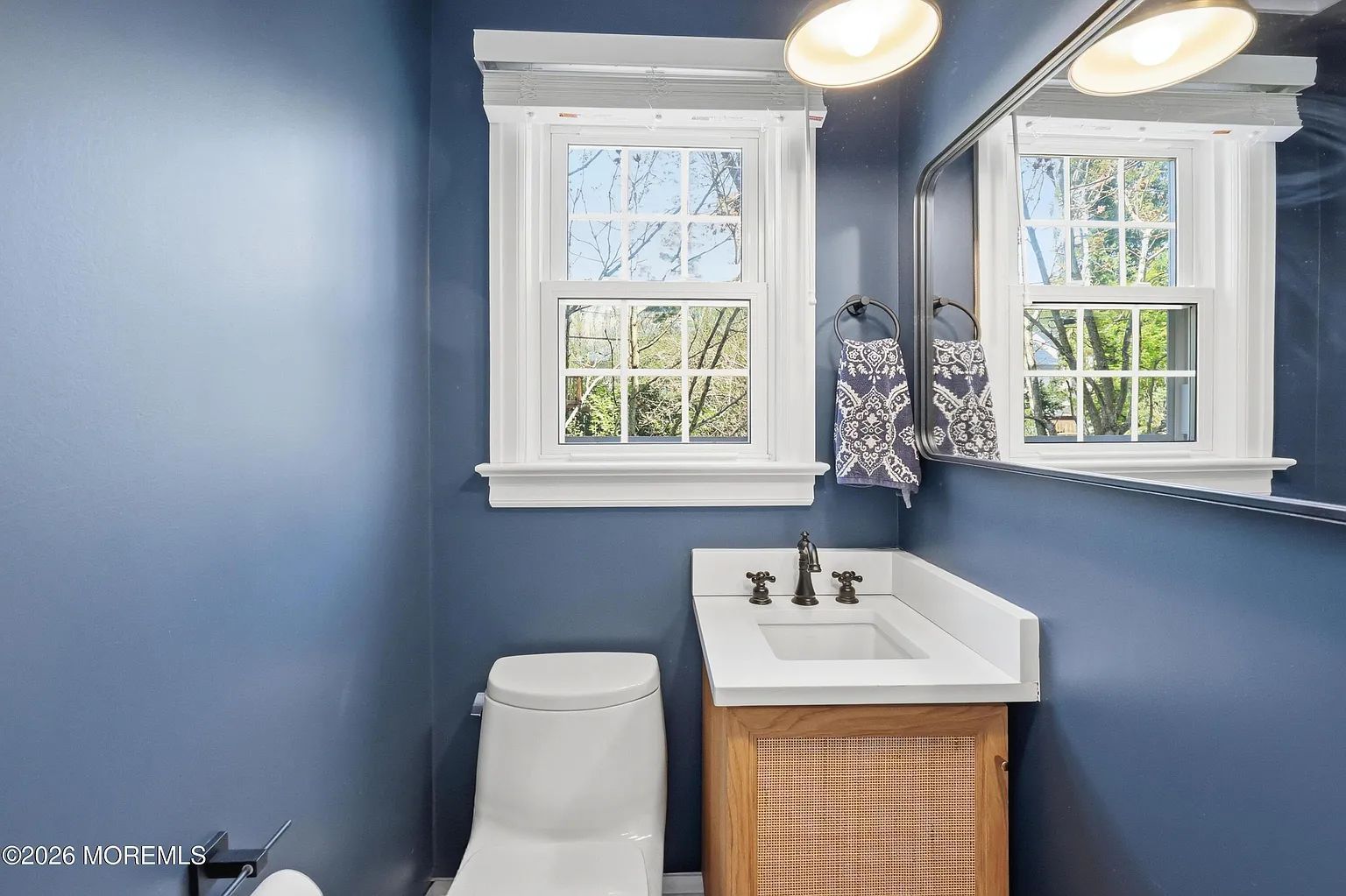 Blue bathroom with white sink, toilet, mirror, and two windows with patterned curtains
