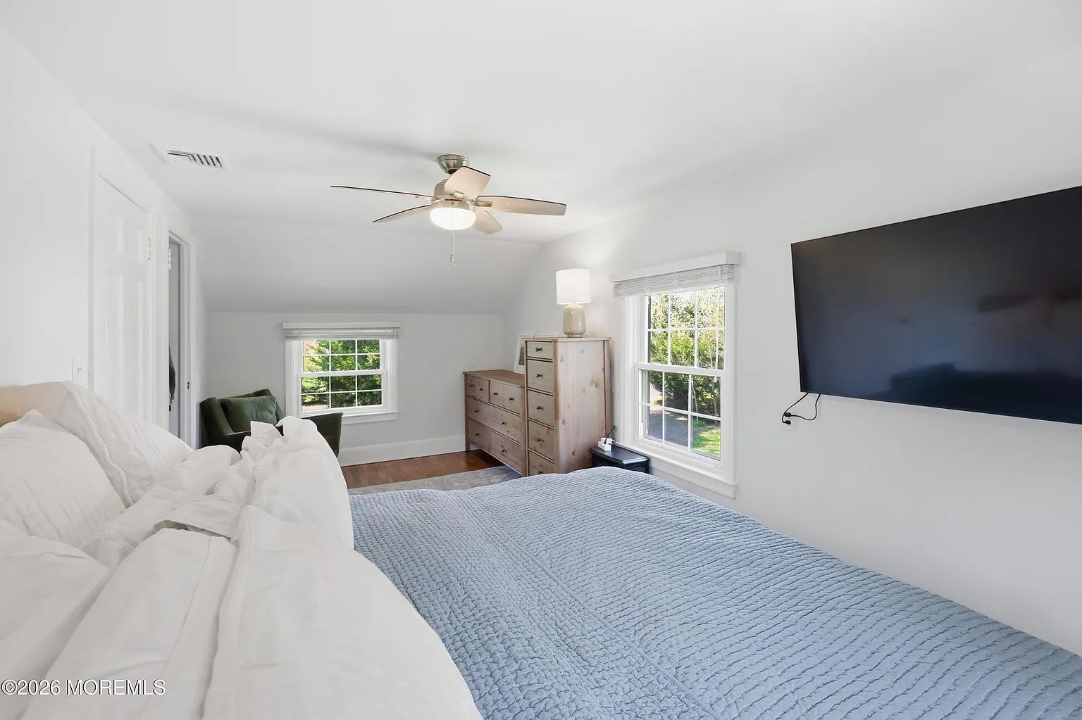 Bright bedroom with white walls, blue bedspread, wall-mounted TV, and two windows with greenery outside