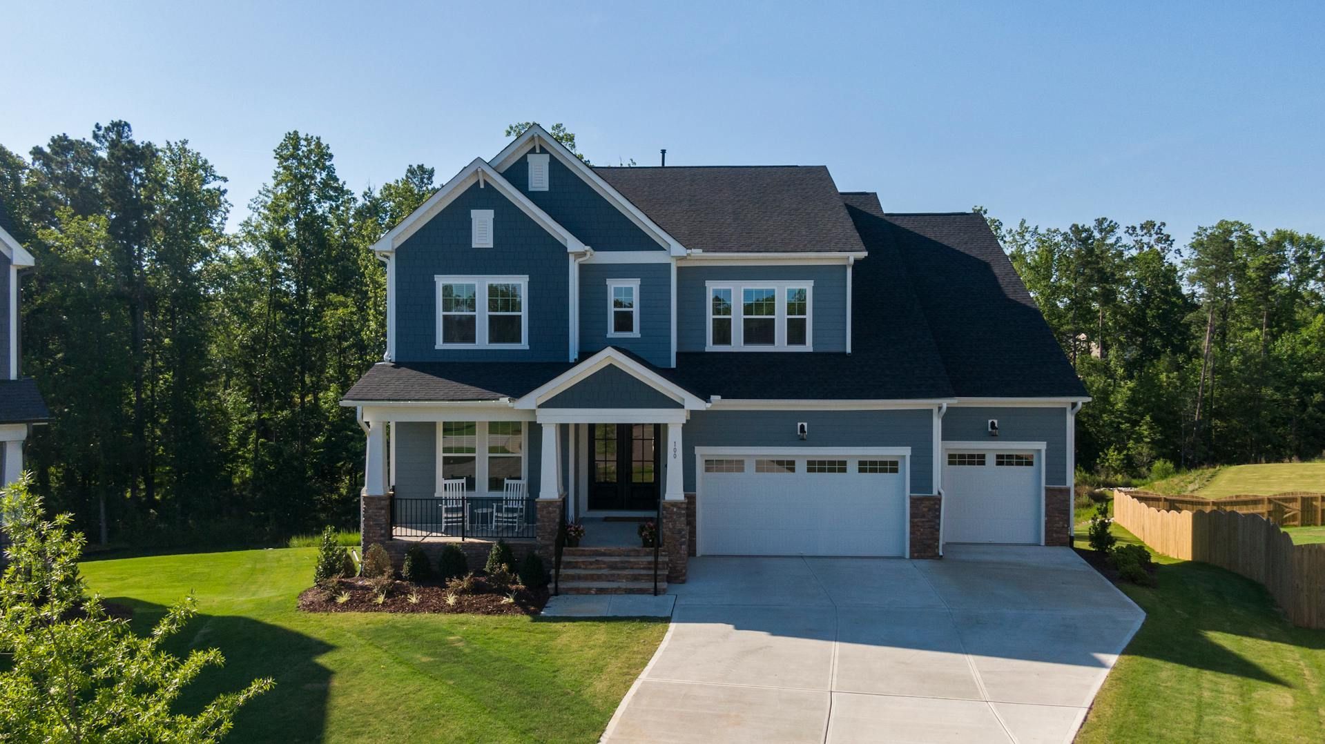 Blue two-story house with attached two-car garage, front porch, and driveway in a wooded suburban setting