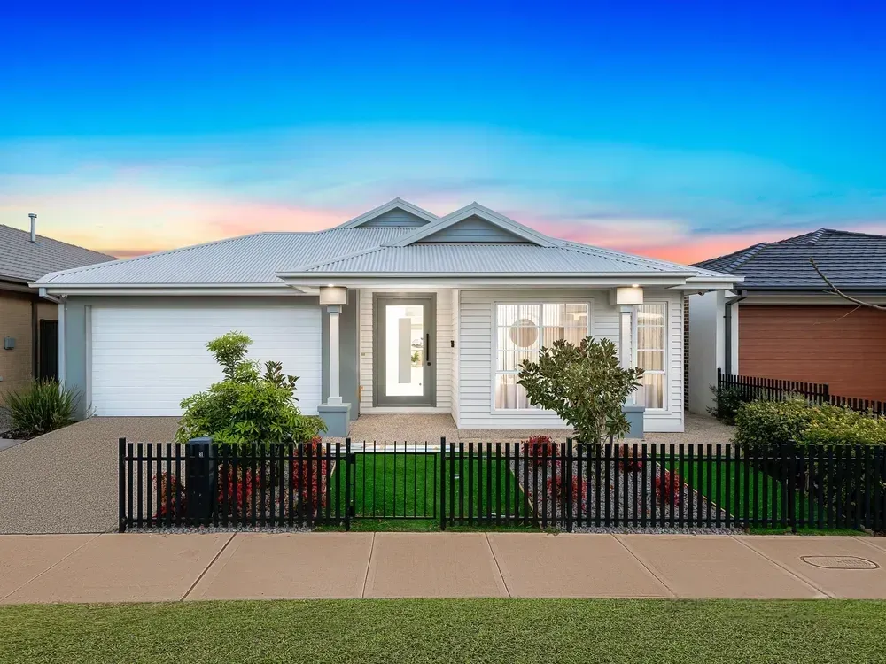 Single-story modern house with a white facade, gray roof, and black picket fence, set against a colorful sunset sky.