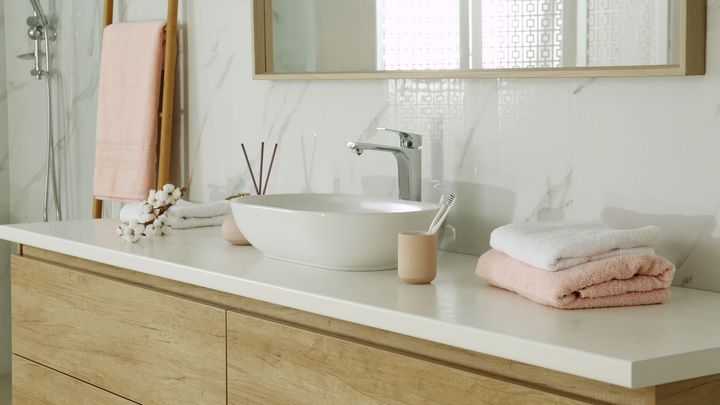 Bathroom sink with a white countertop, wooden vanity, and marble tile. Includes a vessel sink, mirror, towels, and accessories in neutral tones.