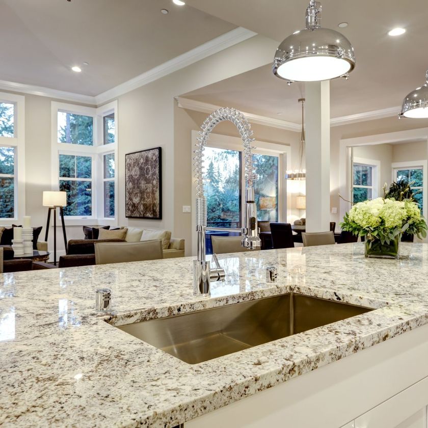 Kitchen island with granite countertop, stainless steel sink, and arched faucet; open to a living room with windows.