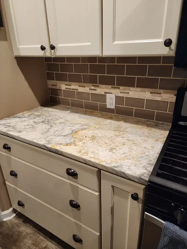 Kitchen undergoing renovation; wooden cabinets, an island, and a black stove are present amidst construction debris. Light gray flooring and white walls are visible.