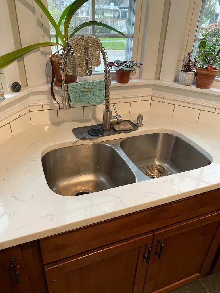 Kitchen sink with stainless steel basins, stainless faucet, and a white countertop with tile backsplash. Wooden cabinets are below.