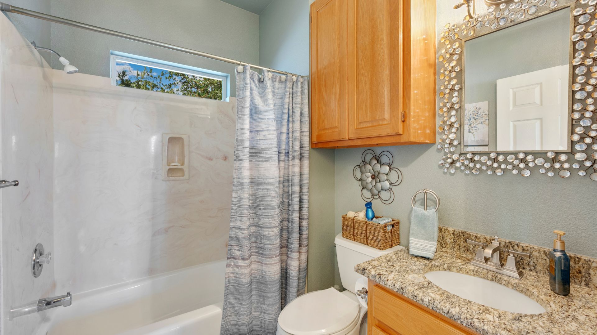 Bathroom with a white bathtub, light blue walls, and a wood cabinet above the toilet. A decorative mirror and a window are also visible.