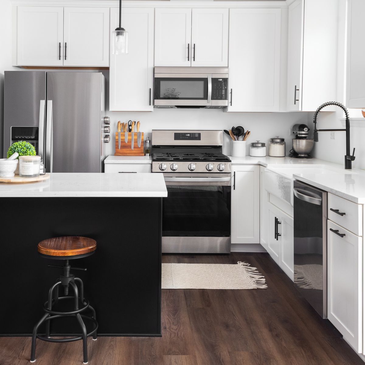 Modern white kitchen with a black island, stainless steel appliances, and dark wood floors. A stool sits at the island.