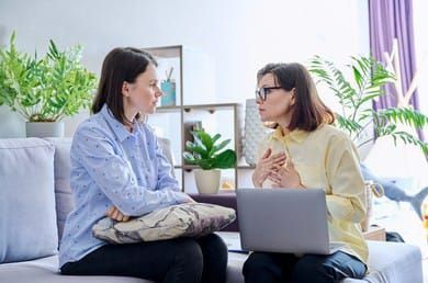 Dos mujeres están sentadas en un sofá con una computadora portátil y hablando entre sí.