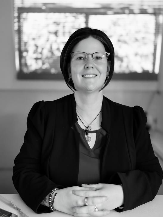 Person with glasses, dark hair, and a dark blazer sitting at a table with hands clasped, smiling at the camera.