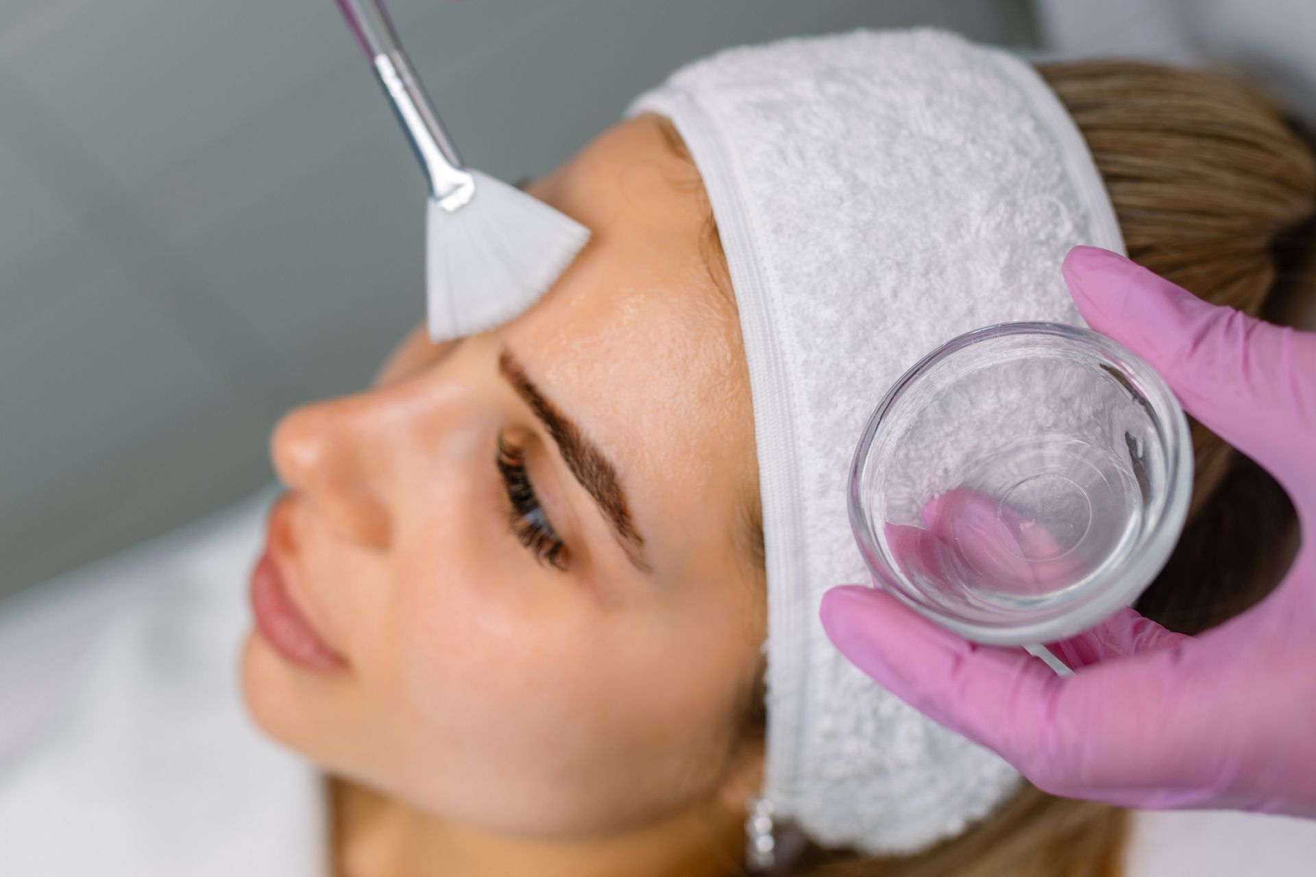 Woman receiving a facial peel treatment; a professional applies liquid with a brush in a clinic setting.