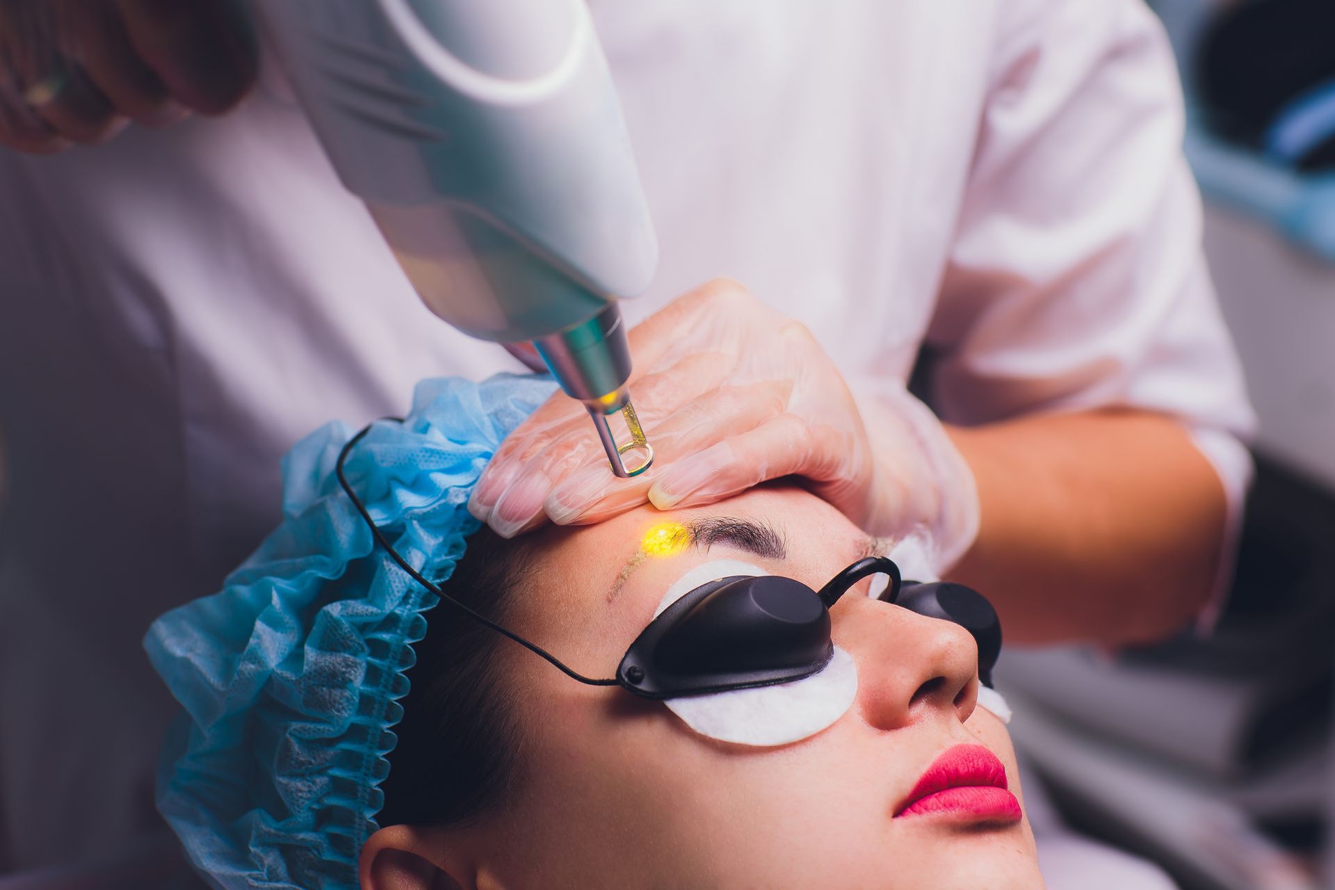 Person receiving laser treatment on their eyebrow, wearing protective eyewear and cap.