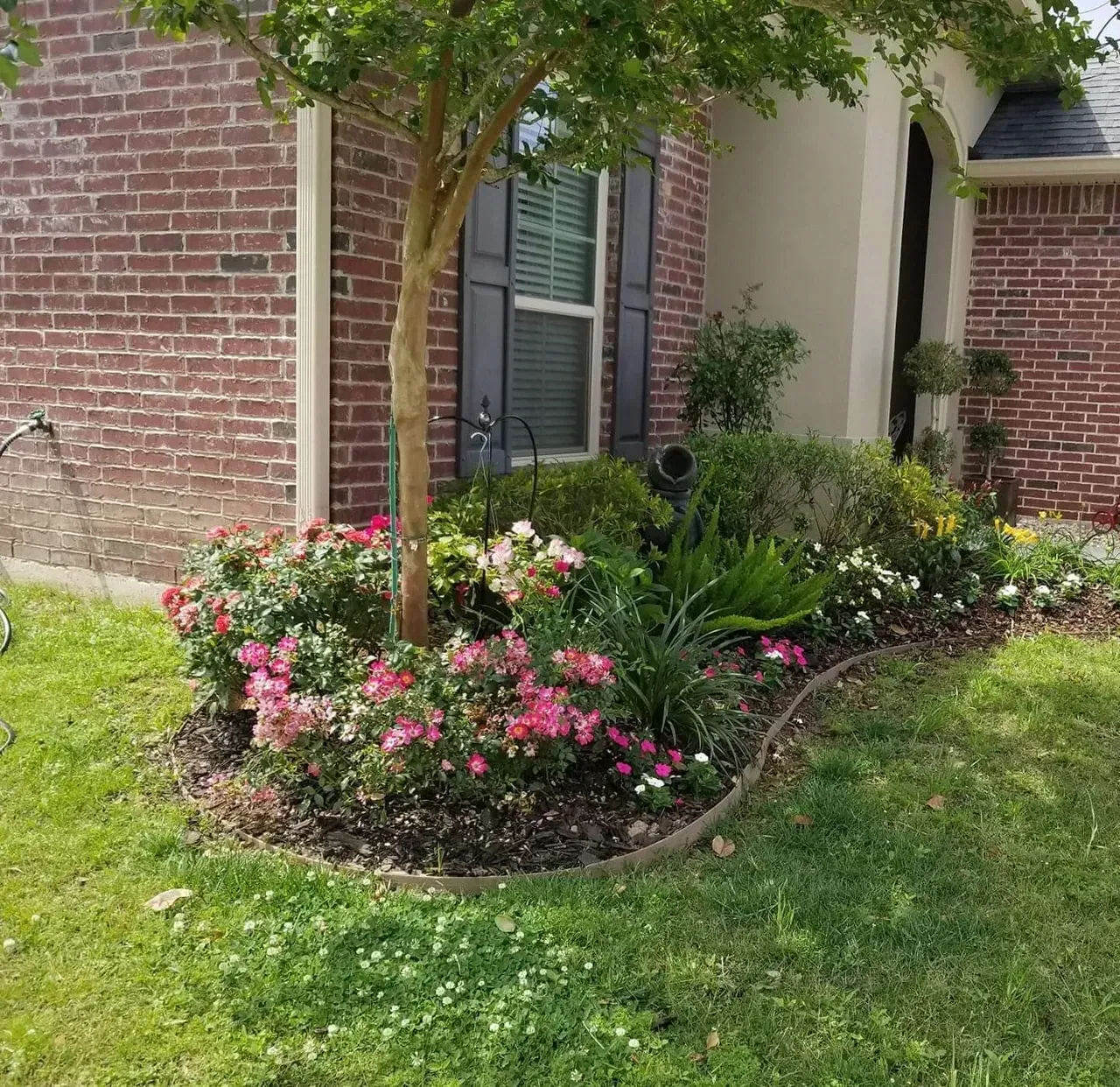 Flowering plants border a brick house; a tree is in the center.