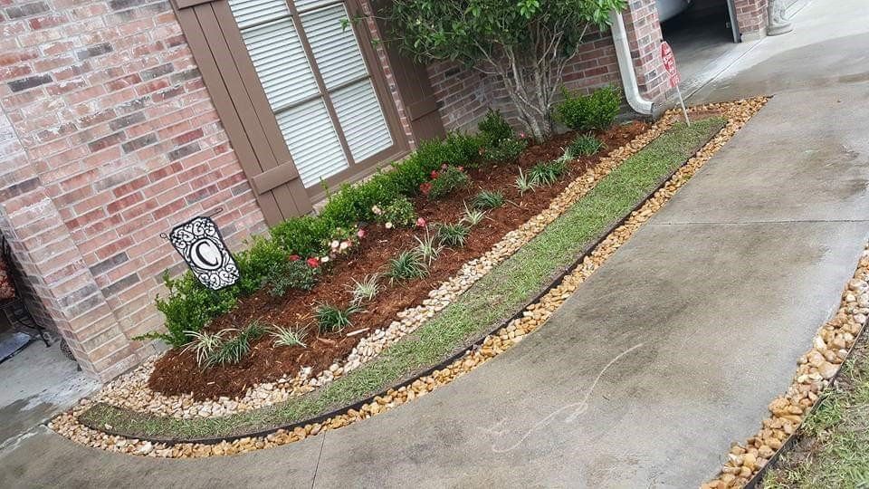 Landscaped front yard bed with mulch, plants, and rocks bordering a driveway. Brick house visible.
