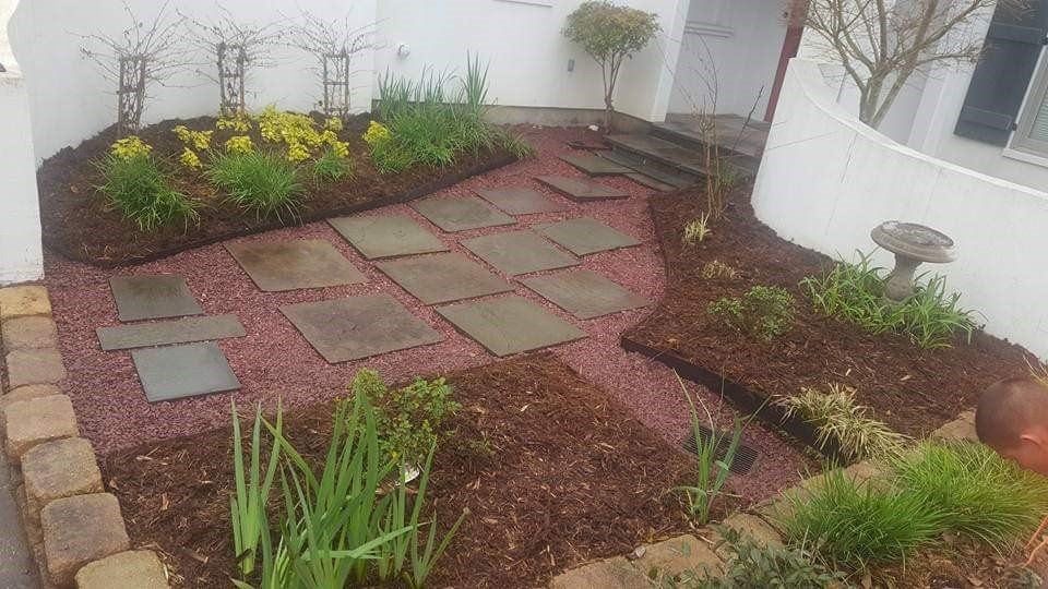 Garden bed with stone path, mulched with red gravel and brown bark, bordered by brick and white walls.