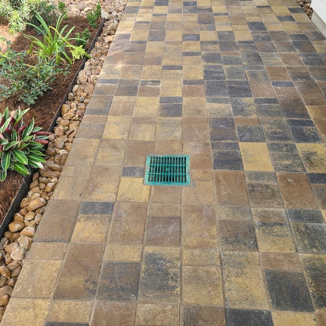 Brick walkway with green drain in the center, bordered by plants and tan stones.