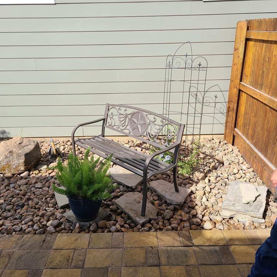 Metal bench in a rock garden, with a potted plant, next to a wooden fence.