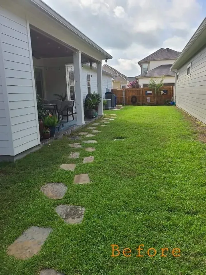 Backyard with stone path, lawn, fence, and houses under a cloudy sky.